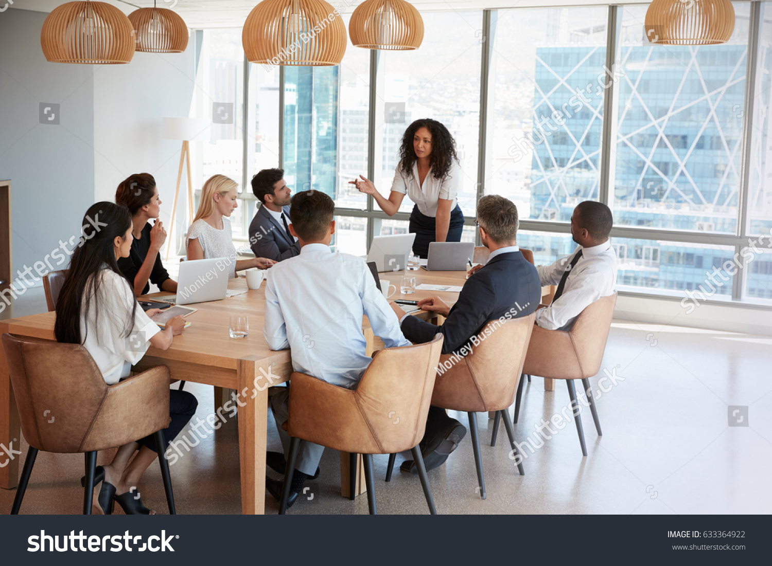 stock-photo-businesswoman-stands-to-address-meeting-around-board-table-633364922